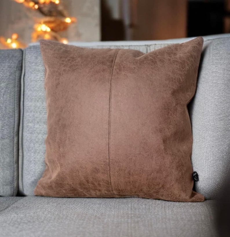 Peut inclure: Un coussin en daim brun avec une couture discr&egrave;te au centre. Le coussin est pos&eacute; sur un canap&eacute; gris clair.