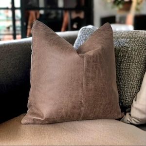 Peut inclure: Un coussin en cuir brun avec un aspect vieilli subtil. Le coussin est pos&eacute; sur un canap&eacute; avec d'autres coussins.