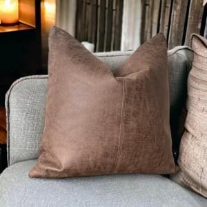 Peut inclure: Un coussin en simili cuir marron avec une surface textur&eacute;e subtile. Le coussin a une couture au centre et est pr&eacute;sent&eacute; sur un canap&eacute; gris.