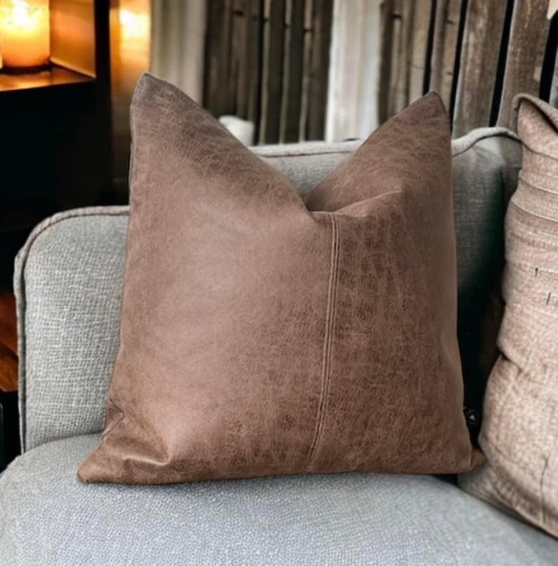 Peut inclure: Un coussin en simili cuir marron avec une surface textur&eacute;e subtile. Le coussin a une couture au centre et est pr&eacute;sent&eacute; sur un canap&eacute; gris.