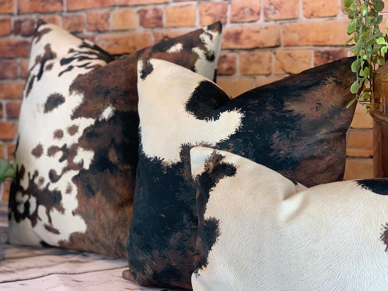 May include: Close-up of three decorative throw pillows with a cowhide pattern. The pillows feature a mix of white, brown, and black patches, creating a rustic and textured appearance. The pillows are arranged in a layered composition, suggesting a cozy home decor theme.