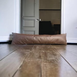 May include: A brown leather doorstop in the shape of a long pillow. The doorstop is lying on a wooden floor in front of a closed white door.