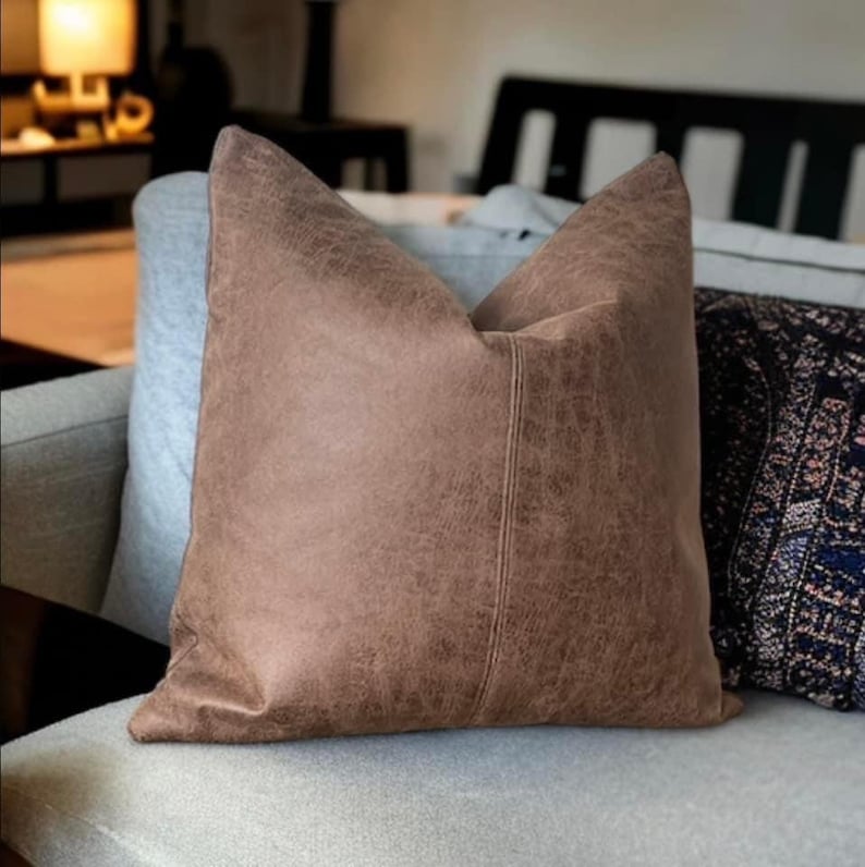 Peut inclure: Un coussin en cuir marron avec un aspect vieilli subtil. Le coussin a une couture au centre et est plac&eacute; sur un canap&eacute; gris clair.