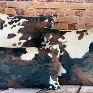 May include: A set of four decorative throw pillows with a cowhide pattern. The pillows feature a mix of white, brown, and black colors. The pillows are arranged in a group, showcasing their different sizes and shapes. The pillows are placed on a wooden surface.