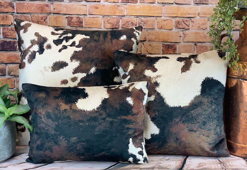 May include: A set of four decorative throw pillows with a cowhide pattern. The pillows feature a mix of white, brown, and black colors. The pillows are arranged in a group, showcasing their different sizes and shapes. The pillows are placed on a wooden surface.