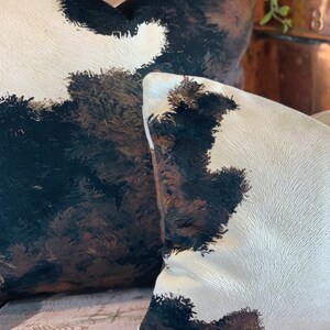 May include: Two decorative throw pillows with a cowhide pattern. The pillows feature a cream-colored background with large patches of black, brown, and tan, creating a rustic and textured appearance. The pillows are placed on a wooden surface.