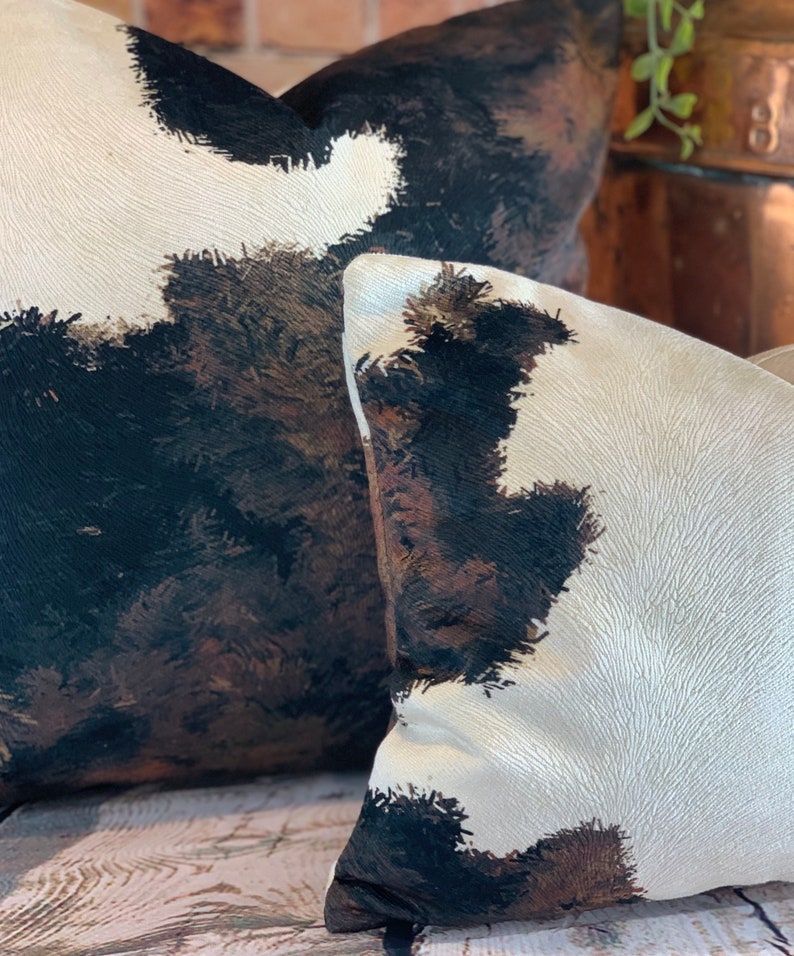 May include: Two decorative throw pillows with a cowhide pattern. The pillows feature a cream-colored background with large patches of black, brown, and tan, creating a rustic and textured appearance. The pillows are placed on a wooden surface.