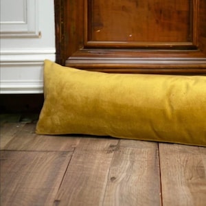 May include: A long, mustard yellow velvet bolster pillow. The pillow is lying on a wooden floor.