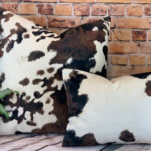 May include: Two decorative throw pillows with a cowhide pattern. The pillows feature a white background with brown and black splotches. The pillows are different sizes and are placed in front of a brick wall.