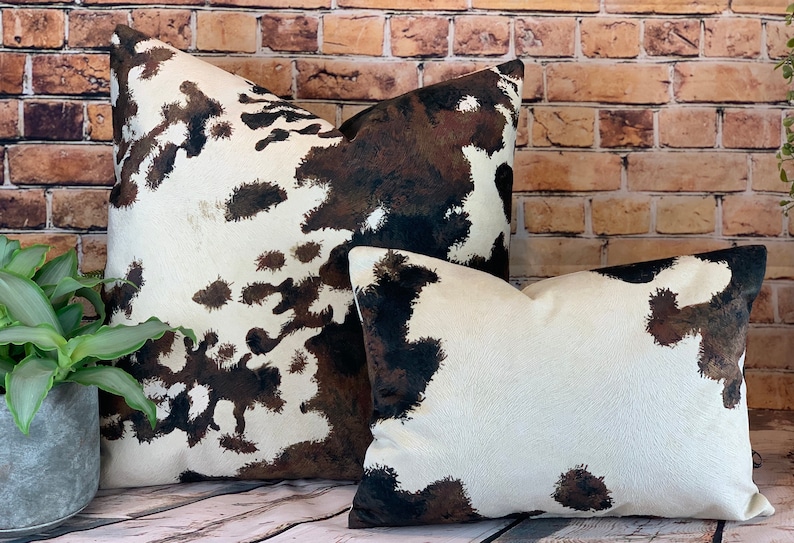 May include: Two decorative throw pillows with a cowhide pattern. The pillows feature a white background with brown and black splotches. The pillows are different sizes and are placed in front of a brick wall.