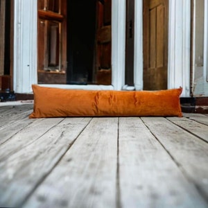 May include: A long, orange velvet bolster pillow sits on a wooden floor.