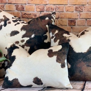 May include: Four decorative throw pillows with a cowhide pattern. The pillows feature a white background with brown and black splotches. The pillows are arranged in front of a brick wall and a potted plant.