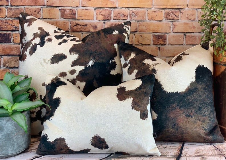 May include: Four decorative throw pillows with a cowhide pattern. The pillows feature a white background with brown and black splotches. The pillows are arranged in front of a brick wall and a potted plant.
