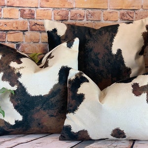 May include: Three decorative throw pillows with a cowhide pattern. The pillows feature a cream-colored background with brown and black splotches, mimicking the look of cowhide. The pillows are arranged in front of a brick wall.