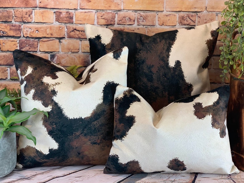 May include: Three decorative throw pillows with a cowhide pattern. The pillows feature a cream-colored background with brown and black splotches, mimicking the look of cowhide. The pillows are arranged in front of a brick wall.