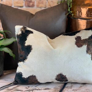 May include: Two decorative throw pillows. The front pillow has a white background with brown and black cowhide-style spots. The second pillow is a solid dark brown. The pillows are on a wooden surface.