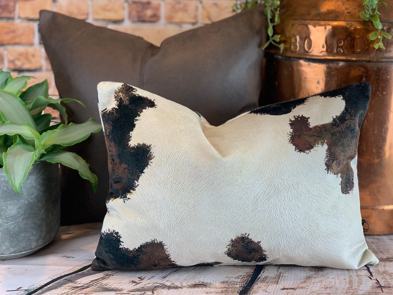 May include: Two decorative throw pillows. The front pillow has a white background with brown and black cowhide-style spots. The second pillow is a solid dark brown. The pillows are on a wooden surface.