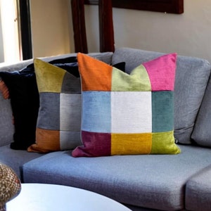 May include: Two decorative throw cushions on a grey sofa. The cushion on the right is a patchwork of orange, green, blue, white, purple, and yellow squares. The cushion on the left is a patchwork of grey, brown, and green squares.