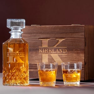 May include: A wooden box with a personalized engraved initial "K" and the name "Kirkland" on the front. The box contains a crystal decanter with a personalized engraved initial "K" and the name "Kirkland" and two matching glasses with the same personalization.