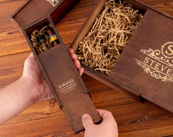 May include: A wooden box with a sliding lid, engraved with the text "DAVID GROOMSMAN 09.10.2023". The box contains a small bottle and a cigar. The box is open and the lid is being held by a hand.