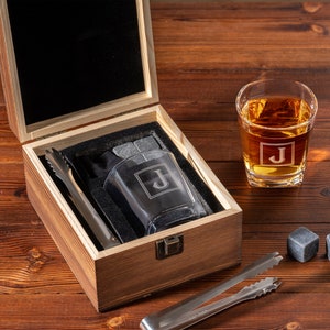 May include: A wooden box with a black interior contains a set of whiskey stones, a pair of tongs, and a whiskey glass. The whiskey glass and stones are engraved with the letter "J".