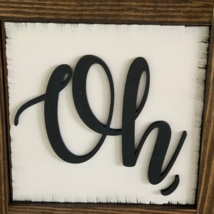 May include: A white wooden sign with a black cutout of the word "Oh" in a cursive font. The sign is framed with a dark brown wood frame.