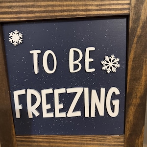 May include: A wooden sign with a blue background and white glitter. The sign says "To Be Freezing" with white snowflakes on either side of the text.