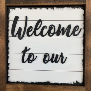 May include: Wooden sign with the words "Welcome to our" in black script. The sign has a white background with horizontal wooden slats and a dark brown frame. The edges of the white background have a distressed, worn look.