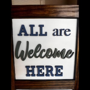 May include: Wooden framed sign with the words "ALL are Welcome HERE" in navy blue and dark green 3D lettering on a white background. The frame is a dark brown color.
