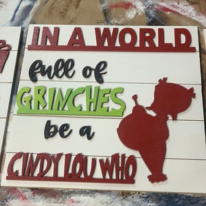 May include: A white wooden sign with the text "In a world full of Grinches be a Cindy Lou Who" in red, black, and green letters. A red silhouette of a person is on the right side of the sign.