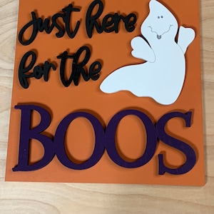 May include: Orange wooden sign with the text "Just here for the boos" in black lettering. A white ghost with a smiling face is above the text. The word "boos" is in large purple letters.