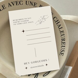 May include: A beige postcard with black text that reads "Remember to say you're enough to your love ones."  The postcard also features a line drawing of two hands reaching towards each other with stars in between.  The bottom of the postcard reads "Hey Gorgeous! @yoursocialmedia"