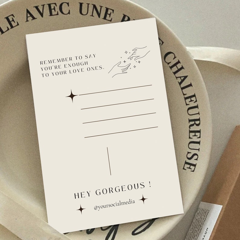 May include: A beige postcard with black text that reads "Remember to say you're enough to your love ones."  The postcard also features a line drawing of two hands reaching towards each other with stars in between.  The bottom of the postcard reads "Hey Gorgeous! @yoursocialmedia"