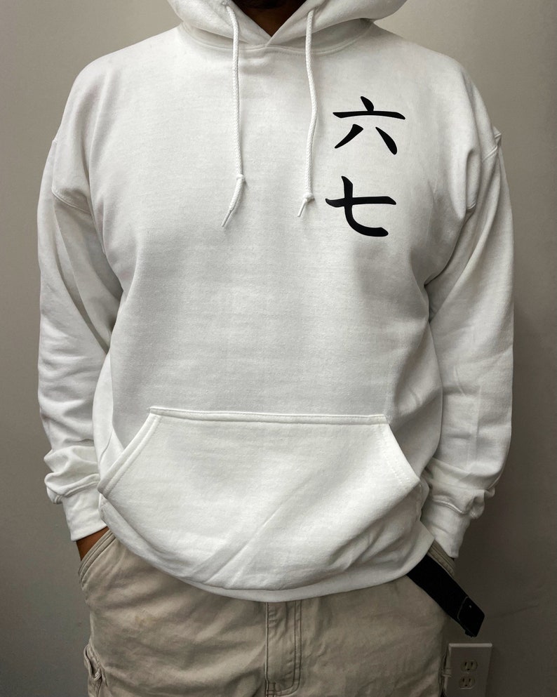 May include: White hooded sweatshirt with a front pocket and drawstring. The sweatshirt features black Japanese characters on the upper left chest. The person is wearing khaki pants and a black belt.