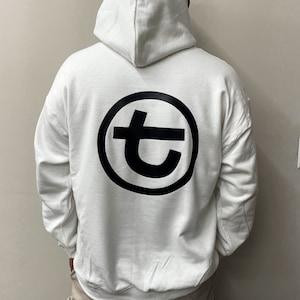 May include: White hooded sweatshirt with a large black circular logo on the back. The logo features a stylized letter "t" inside a circle. The sweatshirt has a drawstring hood and long sleeves.