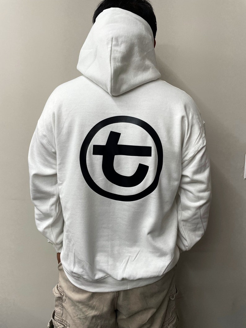 May include: White hooded sweatshirt with a large black circular logo on the back. The logo features a stylized letter "t" inside a circle. The sweatshirt has a drawstring hood and long sleeves.