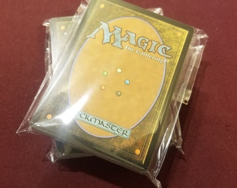 May include: Sealed decks of Magic: The Gathering trading cards. The card backs feature a gold and brown design with the word "Magic" in a stylized font. The decks are packaged in clear plastic.