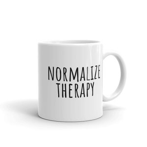 May include: White ceramic mug with the words "NORMALIZE THERAPY" printed in black, sans-serif font. The mug has a curved handle and a glossy finish. The mug is a standard size for coffee or tea.