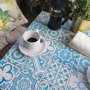 May include: A white ceramic coffee cup and saucer on a table with blue and white patterned tiles. A glass French press filled with coffee is also on the table.