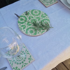 May include: A set of four green and white ceramic coasters with a geometric pattern. The coasters are on a white tablecloth with a glass of water and a plate on the table.