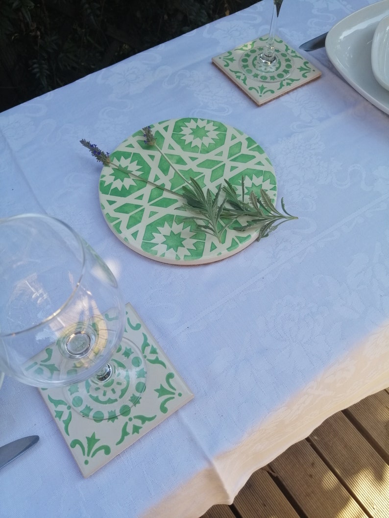 May include: A set of four green and white ceramic coasters with a geometric pattern. The coasters are on a white tablecloth with a glass of water and a plate on the table.
