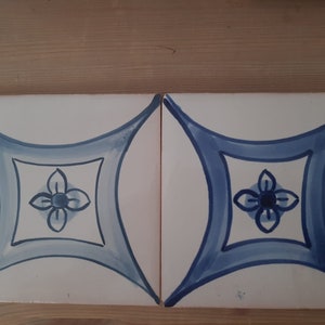 May include: Two white ceramic tiles with a blue and white floral design. The design features a stylised flower with four petals in the centre of each tile.