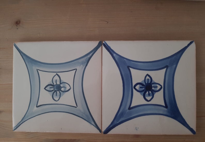 May include: Two white ceramic tiles with a blue and white floral design. The design features a stylised flower with four petals in the centre of each tile.