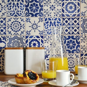 Peut inclure: Un comptoir de cuisine avec un dosseret en carrelage &agrave; motifs bleus et blancs. Il y a deux pots en c&eacute;ramique blanche avec des couvercles en bois, un pichet en verre rempli de jus d'orange, deux verres de jus d'orange et deux tasses &agrave; caf&eacute; blanches avec des soucoupes. Il y a deux p&acirc;tisseries sur une assiette blanche.