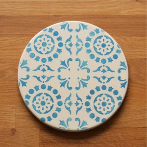 May include: A white ceramic coaster with a blue and white geometric pattern. The coaster features a repeating design of stars, swirls, and circles.
