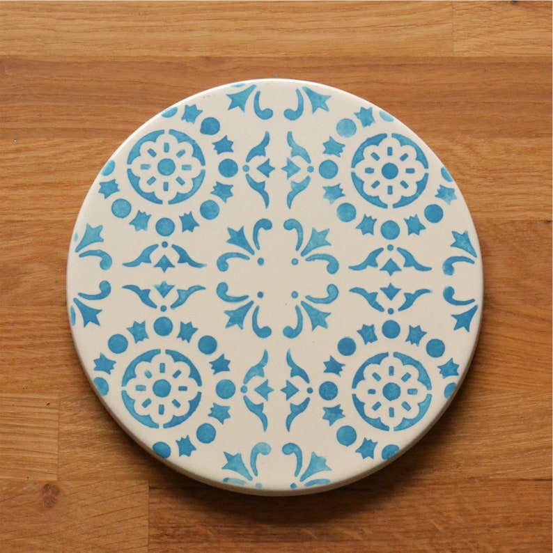 May include: A white ceramic coaster with a blue and white geometric pattern. The coaster features a repeating design of stars, swirls, and circles.