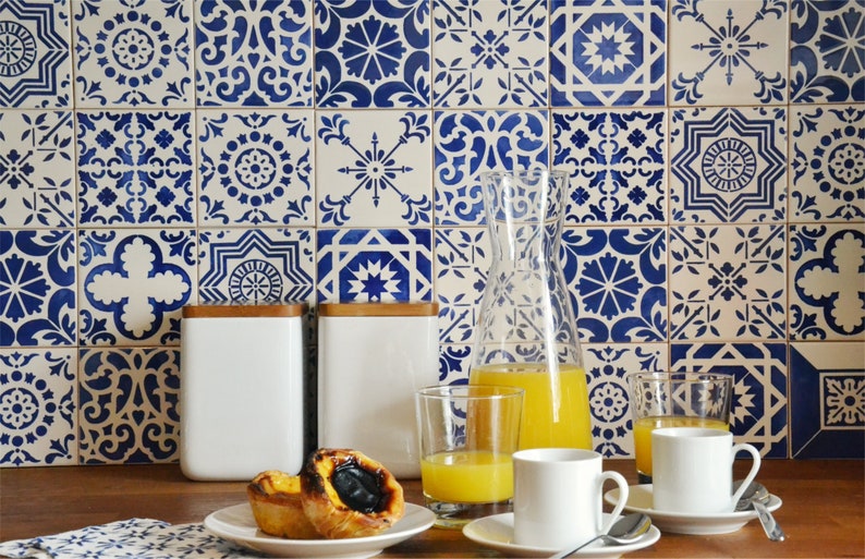 May include: A kitchen counter with a blue and white patterned tile backsplash. There are two white ceramic canisters with wooden lids, a glass pitcher filled with orange juice, two glasses of orange juice, and two white coffee cups with saucers. There are two pastries on a white plate.