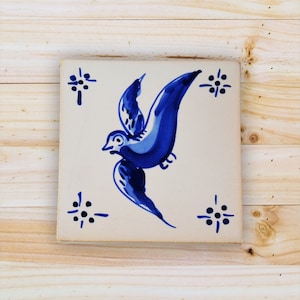 May include: A white ceramic tile featuring a blue bird design. The bird is depicted in flight with its wings outstretched. The tile has a white background with blue floral patterns in the corners.