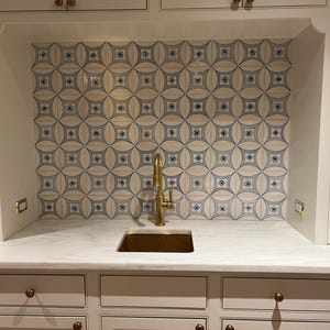 Op de afbeelding: Een keuken- of barruimte met witte kasten, een marmeren aanrechtblad en een messing kraan en spoelbak. De achterwand is voorzien van tegels met een crème- en blauw patroon. Gouden hardware accentueert de kasten.