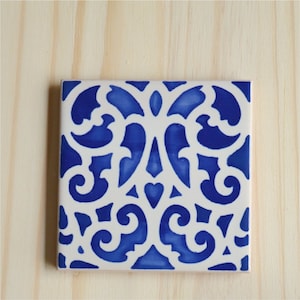May include: A white ceramic tile with a blue and white intricate pattern. The pattern is a repeating design of swirls and curves.